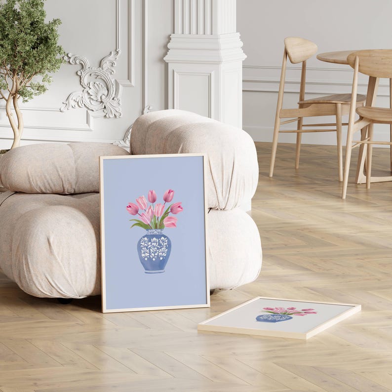 May include: Two framed art prints featuring a floral still life. The artwork depicts pink tulips in a blue and white vase against a light blue background. The prints are displayed on a wooden floor near a beige sofa.