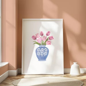 May include: A framed print featuring a blue and white decorative vase filled with pink tulips. The vase has a detailed floral design. The artwork is displayed on a white background, leaning against a peach-colored wall. A white vase sits on the floor.
