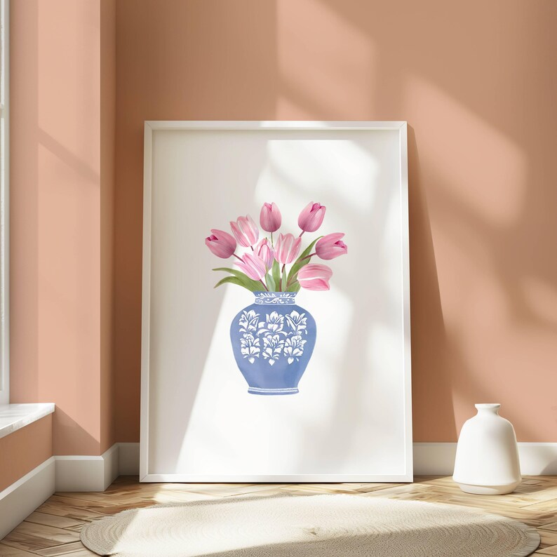 May include: A framed print featuring a blue and white decorative vase filled with pink tulips. The vase has a detailed floral design. The artwork is displayed on a white background, leaning against a peach-colored wall. A white vase sits on the floor.