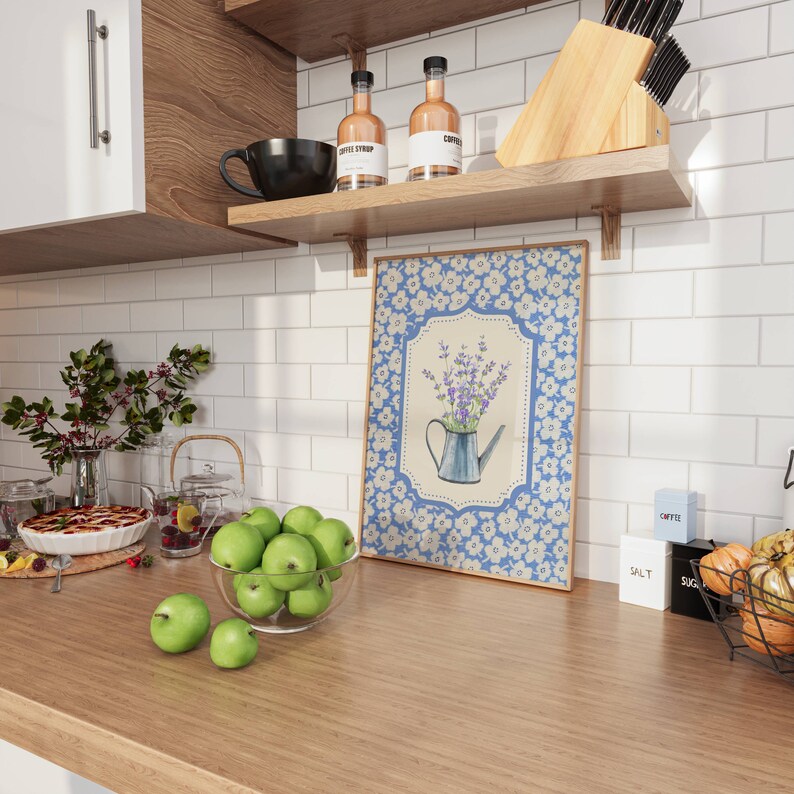 May include: A kitchen scene with a wooden countertop, white tile backsplash, and wooden shelves. A framed floral print, a bowl of green apples, and various kitchen items are displayed. Coffee syrup bottles and a black mug are on the shelves.