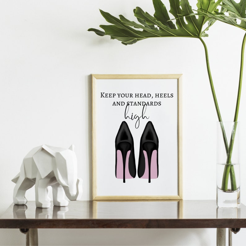 May include: A framed print with the text "Keep your head, heels and standards high" above an illustration of black high heels with pink soles. A white geometric elephant sculpture and a vase with green leaves are on a dark brown surface.