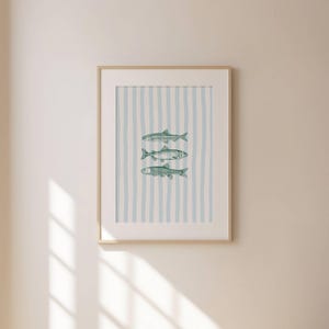 May include: Framed art print featuring three stylized fish in shades of green. The background has vertical stripes of light blue and off-white. The artwork is displayed in a light wood frame.