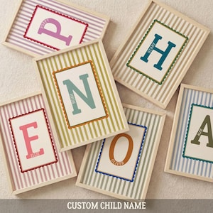 May include: Framed custom child name art prints. Each print features a large initial letter in a bold color, with the child's name below. The background has a striped pattern in various pastel colors. The frames are light wood.