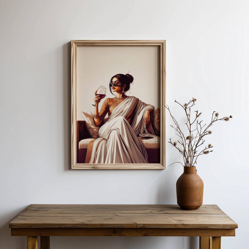 Puede incluir: Impresi&oacute;n art&iacute;stica enmarcada de una mujer con un sari blanco, sosteniendo una copa de vino tinto. La obra presenta a una mujer con cabello oscuro y gafas de sol, sentada en un sof&aacute;. La impresi&oacute;n est&aacute; en un marco de madera, exhibida en una pared blanca sobre una mesa de madera.