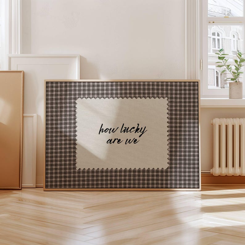May include: A framed wall art piece with a brown and white checkered pattern border. The center features a light beige fabric with the handwritten text "how lucky are we" in black cursive font. The frame is made of wood.
