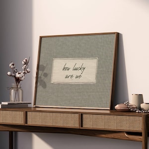 May include: A framed print with a green and white gingham pattern and the text "how lucky are we." The frame is a dark wood. The print is displayed on a wooden console table with decorative items, including a vase of cotton stems.