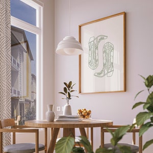 May include: A framed print showing a green line drawing of two cowboy boots. The artwork is displayed in a room with a round wooden table, chairs, and a window with a view of a building. A white pendant light hangs above the table.