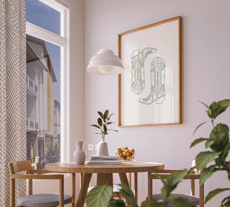 May include: A framed print showing a green line drawing of two cowboy boots. The artwork is displayed in a room with a round wooden table, chairs, and a window with a view of a building. A white pendant light hangs above the table.
