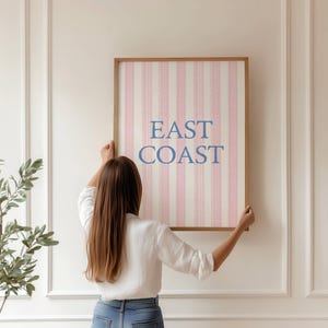 May include: A framed wall art print with the words "EAST COAST" in blue lettering. The artwork features a pink and white striped pattern. The frame is a light brown color. The artwork is being held up by a person.
