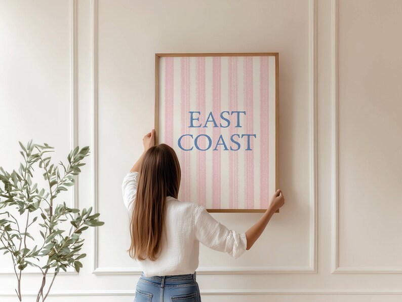May include: A framed wall art print with the words "EAST COAST" in blue lettering. The artwork features a pink and white striped pattern. The frame is a light brown color. The artwork is being held up by a person.