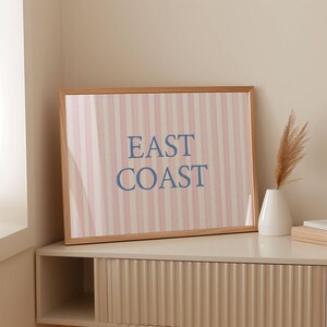 May include: A framed print with the words "EAST COAST" in blue letters. The background features vertical stripes in pink and white. The frame is made of light-colored wood. The print is displayed on a white cabinet.