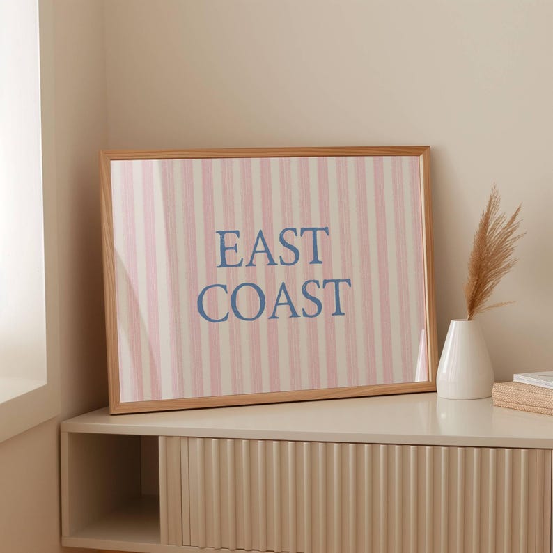 May include: A framed print with the words "EAST COAST" in blue letters. The background features vertical stripes in pink and white. The frame is made of light-colored wood. The print is displayed on a white cabinet.