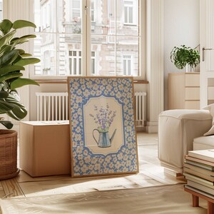 May include: A framed print featuring a blue watering can filled with lavender flowers. The print has a light blue floral border and a beige background. The artwork is displayed in a bright, airy room with a large window and a potted plant.