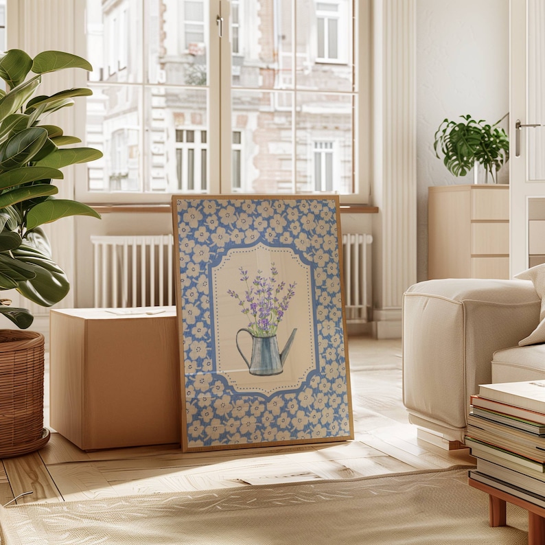 May include: A framed print featuring a blue watering can filled with lavender flowers. The print has a light blue floral border and a beige background. The artwork is displayed in a bright, airy room with a large window and a potted plant.