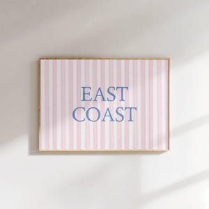 May include: A framed print with a pink and white striped background. The words "EAST COAST" are printed in blue, centered on the print. The frame is a light brown color. The print is hanging on a white wall.