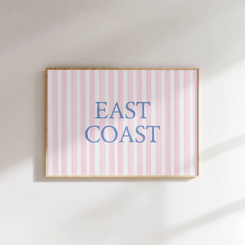 May include: A framed print with a pink and white striped background. The words "EAST COAST" are printed in blue, centered on the print. The frame is a light brown color. The print is hanging on a white wall.
