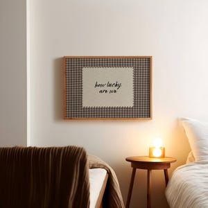 May include: A framed wall art piece with the text "how lucky are we". The artwork features a beige center with the text in black script, surrounded by a brown and white checkered pattern and a wooden frame. The art is hanging on a white wall.