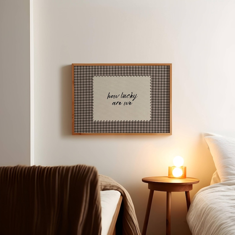 May include: A framed wall art piece with the text "how lucky are we". The artwork features a beige center with the text in black script, surrounded by a brown and white checkered pattern and a wooden frame. The art is hanging on a white wall.