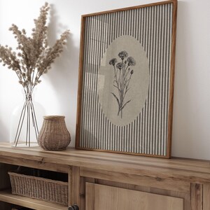 May include: A rectangular framed wall art piece featuring a vertical striped pattern in grey and white with a central beige oval containing a black and white botanical illustration of a cluster of wildflowers with thin stems and textured flower heads.