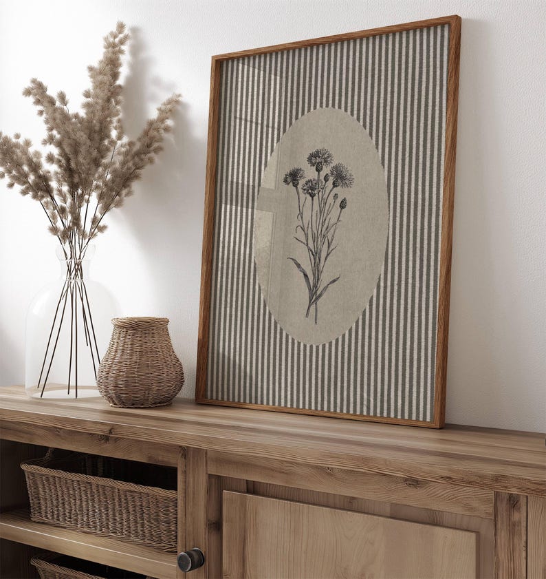 May include: A rectangular framed wall art piece featuring a vertical striped pattern in grey and white with a central beige oval containing a black and white botanical illustration of a cluster of wildflowers with thin stems and textured flower heads.
