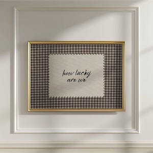 May include: A framed artwork featuring a beige fabric with the phrase "how lucky are we" in black script. The fabric is set against a brown and white checkered background, encased in a gold frame.