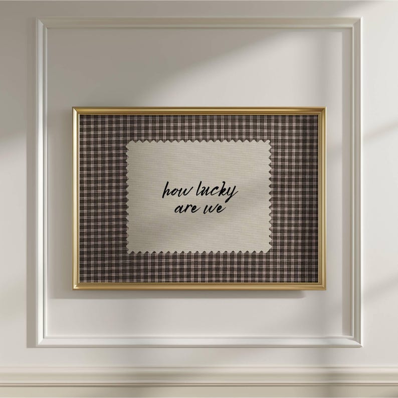 May include: A framed artwork featuring a beige fabric with the phrase "how lucky are we" in black script. The fabric is set against a brown and white checkered background, encased in a gold frame.
