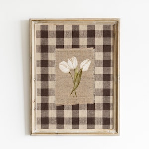 May include: A rectangular wall art piece in a light wood frame featuring a brown and cream buffalo check fabric background. Centered on the fabric is a rectangular piece of hessian displaying a printed illustration of three white tulips with green stems.