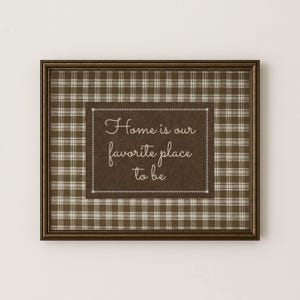 May include: Framed wall art with a brown plaid pattern background and a dark brown center. The center features the quote "Home is our favorite place to be" in elegant script. The frame is a dark brown color.