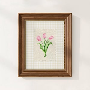 Pode incluir: Uma pe&ccedil;a de arte de parede emoldurada com uma aguarela de tr&ecirc;s t&uacute;lipas cor-de-rosa com caules e folhas verdes sobre papel texturado branco sujo. A obra est&aacute; colocada sobre um fundo axadrezado bege claro e branco numa moldura de madeira.