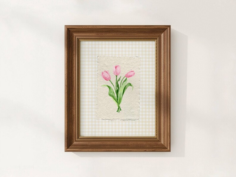 Pode incluir: Uma pe&ccedil;a de arte de parede emoldurada com uma aguarela de tr&ecirc;s t&uacute;lipas cor-de-rosa com caules e folhas verdes sobre papel texturado branco sujo. A obra est&aacute; colocada sobre um fundo axadrezado bege claro e branco numa moldura de madeira.