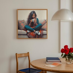 May include: A framed art print shows a person seated cross-legged on a sofa, holding a mug. The person wears teal clothing and white trainers. The sofa has floral cushions and a red patterned blanket. The artwork is in a room with a wooden chair and table.