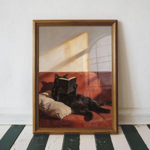 May include: Framed artwork featuring a black cat reclining on a red sofa, engrossed in a book. The cat rests on a cream-colored pillow. The artwork is in a gold-colored frame, set against a white wall and a black and white striped floor.