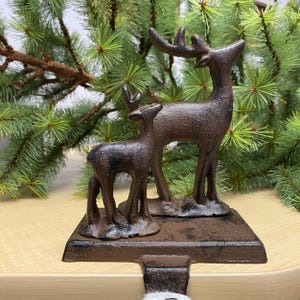 May include: A rustic brown cast iron stocking holder featuring a standing deer and a fawn. The holder has a distressed finish and is perfect for hanging stockings on a fireplace mantel.