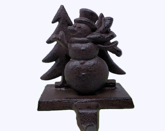 May include: A cast iron stocking holder featuring a snowman with a hat and scarf, standing next to a Christmas tree. The holder is painted in a dark brown color.