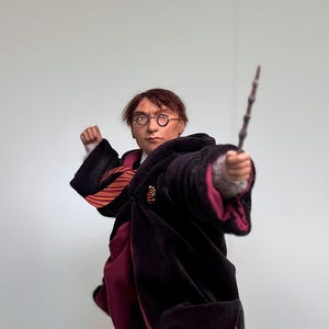 以下が含まれることがあります： 黒地にマルーンの裏地が付いたローブを着た若い魔法使いの細かなフィギュア。赤と金のストライプのネクタイと丸い眼鏡をかけています。彼は右手に杖を持ち、本が置かれた木製の台座の上に立っています。