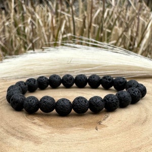 Op de afbeelding: Een zwarte armband van lavastenen kralen. De armband is gemaakt van kleine, ronde kralen die allemaal dezelfde grootte hebben. De kralen zijn aan elkaar geregen op een zwart elastisch koord.