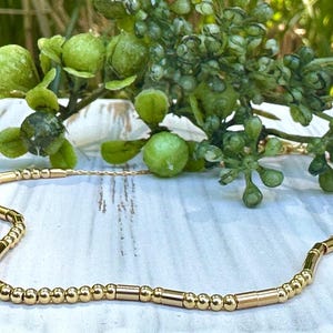 May include: A gold necklace featuring small round beads and longer, cylindrical beads. The necklace is displayed on a white wooden surface.
