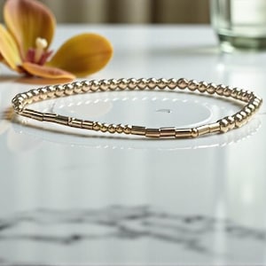 Puede incluir: Una pulsera delicada compuesta por pequeñas cuentas esféricas de color dorado metálico intercaladas con cuentas tubulares cilíndricas cortas de color dorado, dispuestas en un patrón circular continuo.