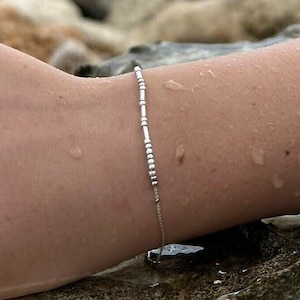 Könnte beinhalten: Ein silbernes Armband mit Perlen und einer zarten Kette. Das Armband wird an einem Handgelenk mit Wassertropfen auf der Haut getragen.