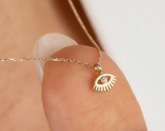 May include: A gold chain necklace with a small eye pendant featuring a single diamond in the centre. The eye is stylised with lashes.