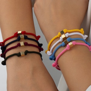 Peut inclure: Deux poignets portant plusieurs bracelets en corde de couleur avec des breloques carrées dorées. Les bracelets sont en rouge, bleu marine, bordeaux, vert foncé, jaune, blanc, bleu clair et rose.