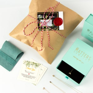 May include: A light green jewelry box with a gold logo that says "Matters Jewelry" and the tagline "What really matters is you." The box is open and contains a gold necklace with a crescent moon pendant. The box is sitting on a white surface next to a brown paper package tied with red and white twine. There is a small green velvet pouch and a thank you card with a floral design.