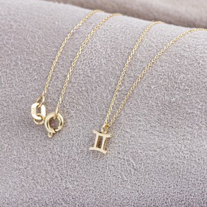 May include: A gold chain necklace with a Gemini zodiac sign pendant. The necklace is on a light gray fabric background.