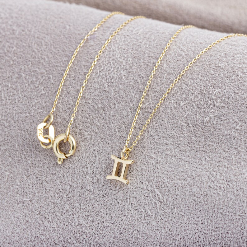 May include: A gold chain necklace with a Gemini zodiac sign pendant. The necklace is on a light gray fabric background.