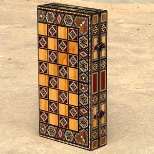 Puede incluir: Un juego de ajedrez de madera cerrado y vertical con intrincados diseños incrustados. El tablero de ajedrez es visible, con cuadrados claros y oscuros alternados. Los lados y los bordes están decorados con diseños geométricos.