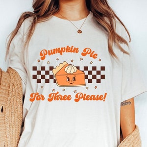 May include: White t-shirt with a retro design featuring a pumpkin pie with a smiling face and the text "Pumpkin Pie For Three Please!"