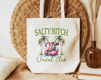 May include: White canvas tote bag featuring the text "Salty Bitch Social Club" in green lettering. A skeleton wearing a sombrero is riding a pink flamingo float in front of two palm trees.