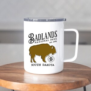 May include: White travel mug featuring the Badlands National Park logo with a bison graphic. The mug has a handle and a silver lid. The text includes "Badlands National Park Est. 1978" and "South Dakota". The mug is on a wooden stand.