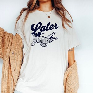 Peut inclure: Un t-shirt blanc avec un graphique d'alligator bleu et le mot "Later" dans une police r&eacute;tro.