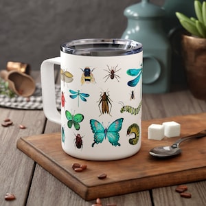 May include: White ceramic mug with a colorful design of various insects and butterflies. The mug has a black handle and a white lid.